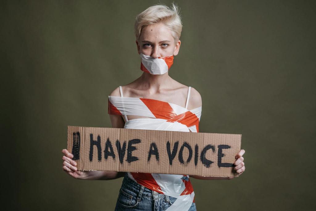 A woman with red tape around her mouth and body silencing her, holding a sign saying 'I have a voice'